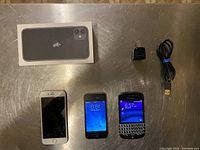 Photo showing the lot with iPhone 11 box, iPhone 6 with broken screen, iPhone 4 powered on, Blackberry Q10 powered on, wall charger, and USB cable arranged on metal surface.