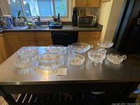 Collection of eleven heavy leaded crystal and glass serving pieces on a metal kitchen island counter. The photo shows various bowls, pedestal dish, and divided dishes with intricate cut designs.