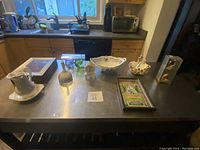 Wide shot of various decor items arranged on a kitchen island, includes porcelain pitchers, decorative bowls, carved figurine, keepsake box, clock, and glass oil lamp among others.