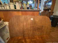 Photo of square oval dining table protection pad with wood grain finish, placed vertically against an indoor counter.