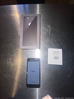 Photo of the iPhone 8 powered on showing 'Hola' on screen, front screen cracked, with original box and lot card.