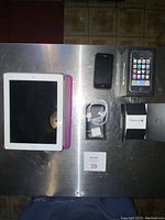 Apple iPad 2 with white bezel in purple case, black iPhone 3GS, iPhone 3GS box, charger and USB cable on metal surface.