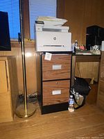 Full view of metal file cabinet with wood grain finish under a desk, showing overall condition and size comparison with nearby floor lamp and printer.