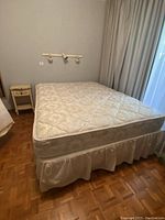 Full view of the metal queen size bed frame with mattress and optional bed skirt (not included). The bed is set up in a room with parquet flooring and has a nightstand beside it.