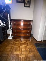 Front view of small solid wood dresser showing four drawers and brass hardware on parquet floor near wall.
