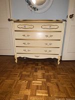 Front view of small vintage dresser showing cream finish with gold decorative accents and ornate drawer handles.