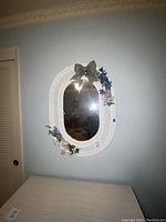 Front view of the white wicker oval mirror hanging on a light blue wall, showing the full decoration and overall condition.