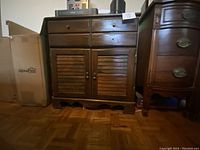 Front view of vintage wooden cabinet with two drawers, one drawer pull missing from each drawer, and two louvered doors below, standing on parquet floor between cardboard box and another wooden furniture piece.