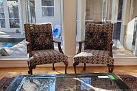 Photo of two upholstered armchairs with decorative pillows placed near large windows on a wooden floor, with a patterned rug underneath. Shows full front view of chairs.