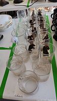 Photo showing twelve tall glasses with black Zodiac sign motifs and four short glasses with gold lily motifs arranged on a table.