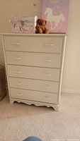 Front view of white chest of drawers with five drawers, showing minor wear and decorative scalloped edge at bottom.