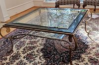 Square coffee table with glass top and marble inlay, iron ornate legs on a patterned carpet.