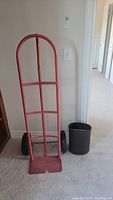 Red metal dolly with wheels and black metal trash can placed beside it, shown in hallway.