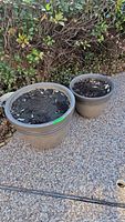 Two flower pots placed on a pebble surface against a green hedge. One larger and one smaller resin planter filled with soil visible.