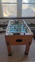 Full view of foosball table showing wooden legs, metal rods with foosball players, playing surface, and window background.