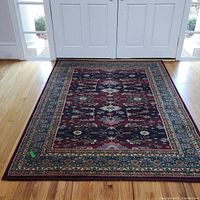 Full view of Kazak rug placed on hardwood floor in front of closed doors. Shows overall color and pattern of rug with central red field and contrasting borders.