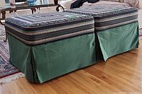 Two square upholstered ottomans with patterned tops and solid green fabric skirt bases on wooden floor near a glass table.