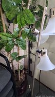 Faux plant with tall branches and green leaves alongside three-light floor lamp with frosted shades.