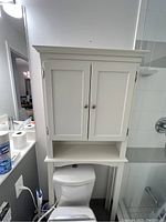 White over-the-toilet bathroom cabinet shown mounted over toilet with two doors and an open shelf below