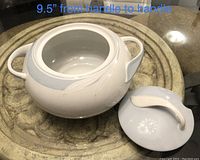 White ceramic soup tureen with lid, showing embossed leaf detail and two handles, placed on a round stone tabletop.