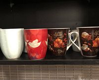 Four coffee mugs on a shelf: white with polka dots, red with white bird pattern, two brown mugs with coffee grinder print
