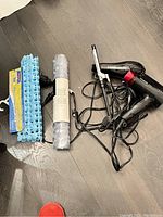 Two black hair dryers, one curling iron, two rolled-up bath mats (one blue, one white/light) on wood floor