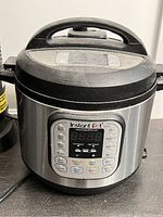 Front-facing photo of Instant Pot electric pressure cooker showing digital control panel with cooking function buttons and stainless steel body with black plastic lid and handle.