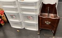 Photo showing two white plastic drawer units and one wooden magazine rack with brass handle and casters on dark flooring.