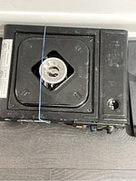 Top view of black portable single burner stove with square burner plate and blue rubber band securing a burner cover, showing signs of usage.