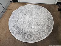 Photo showing the full round rug with visible faded gray floral pattern on white background and scalloped edges.
