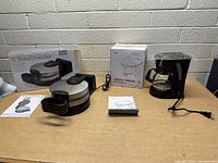 Photo shows boxed Toastmaster waffle maker, unboxed waffle maker, boxed Gevi coffee maker, unboxed coffee maker, instruction manuals, and power cords.