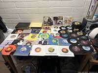 Photo of large assortment of 45 RPM records spread on a table with two album covers propped up in the back.