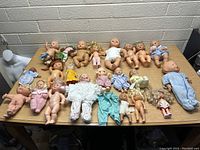 Top-down view of a collection of 24 assorted baby and toddler dolls placed on a wooden table with varying skin tones and clothing.