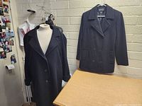 Photo showing both black women's overcoats side by side, one long single-breasted and one shorter double-breasted coat on hangers and mannequin.