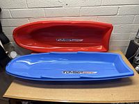 Two Tomser Sport snow sleds stacked on a table, red and blue colors, showing the inner contoured seats and branding.