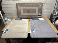 Photo of three memory foam mats: beige and gray Tranquility Collection bath mats with tags visible and a brown Montclair decorative kitchen mat leaning on the wall