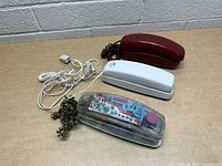 Three vintage phones on a wooden table against a cinder block wall: burgundy, white, and transparent with visible circuitry