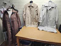 Three women's vintage London Fog coats displayed on hangers and a mannequin: one brown with hood and two beige coats, placed on and behind a table.