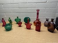 Full view of 10 colorful glass bottles and vases with varied shapes and colors including green, amber, red, and purple.