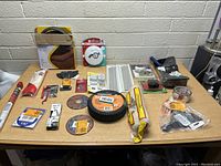 Various assorted tools and hardware items displayed on a table, including vinyl floor trim, saw blades, paint brushes, sandpaper, adhesive tapes, and other repair tools.