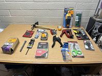 Photo showing assortment of hand tools and accessories laid out on a table including hammer, scissors, clamps, measuring tape, and others.