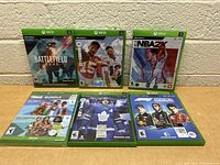 Seven Xbox video game cases arranged on a wooden surface, showing cover art for Battlefield 2042, Madden NFL 22, NBA 2K22, Sims 4 Bundle, NHL 22, and others.