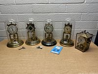 Five vintage mantel clocks showcased on table, four under rounded glass domes with brass bases, one in rectangular lantern-style glass case. Visible winding keys and a Kundo instruction booklet present.