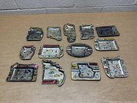 Wide angled photo showing all 13 vintage metal souvenir state ashtrays arranged on a cardboard surface with visible colorful painted designs and various U.S. state shapes.