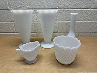 Front view of five vintage milk glass pieces on a table showing hobnail vases, bowl, creamer, and ribbed bud vase.