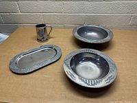 Four-piece metal dishware set displayed on table: two round bowls, rectangular tray, and handled mug with embossed designs.
