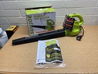 Photo of the Sun Joe leaf blower with box, instruction manuals, and power cord visible, displayed on a table against a brick wall.