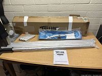 Full view of all components including telescoping poles, snow rake head, and instruction manual on a table in front of a cardboard Tuffiom box.