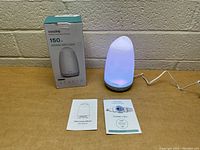 Photo showing diffuser turned on with white light, power cord connected, original box, and manuals on table.