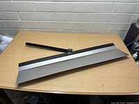 Top angled view showing the bent metal blade attached to the black pivoting handle, placed on a brown table against a white brick wall.